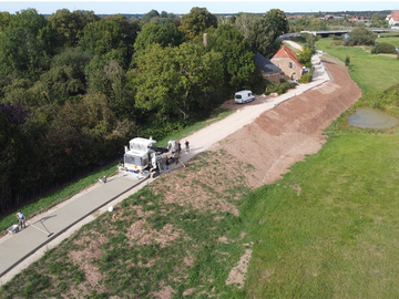 Mit Hilfe eines Betondeckenfertigers wird der Oberbau des Deichverteidigungswegs in kürzester Zeit hergestellt. Auf Höhe der Gebäude am Deich sind die Winkelstützmauer und die mit Deckwerkssteinen befestigte steilere Böschung erkennbar.