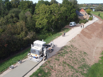 Mit Hilfe eines Betondeckenfertigers wird der Oberbau des Deichverteidigungswegs auf der Deichkrone hergestellt. Auf Höhe der Gebäude am Deich sind die Winkelstützmauer und die mit Deckwerkssteinen befestigte steilere Außenböschung des Deiches erken