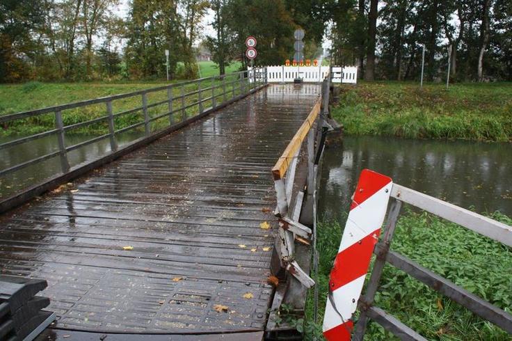 Die beschädigte Brücke im Vordergrund. Unter ihr verläuft der Kanal. Im Hintergrund eine Baumreihe. Absperrungen verhindern das Befahren.