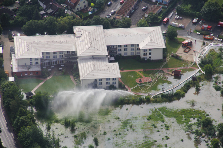 Luftbild: Ein großes Gebäude wird von Hochwasser bedroht. Im Bild wird Pumpen und Wasserwerfern gegen das steigende Wasser gekämpft.