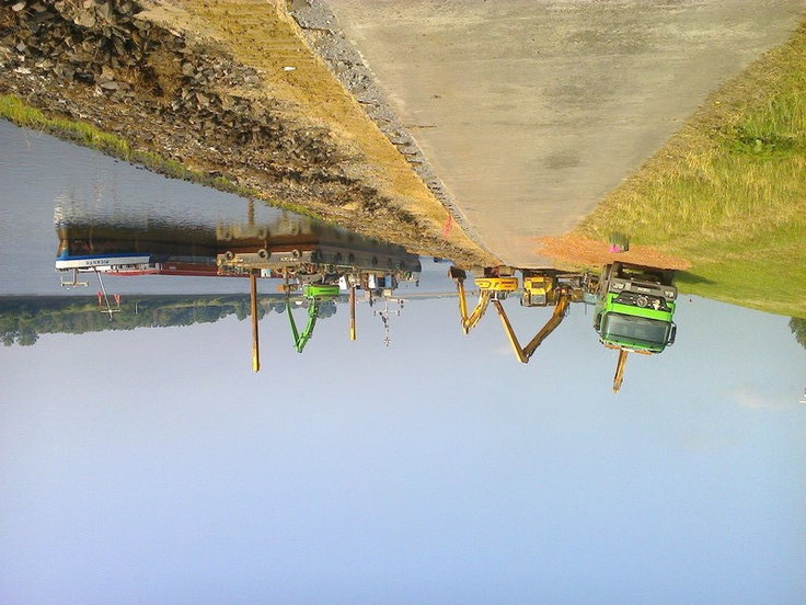 Bild am Deich, links ein Tieflader und Bagger, rechts die Ochtum. Auf ihr liegen zwei Arbeitsboote, von denen aus gemeinsam mit den Geräten an Land an der Herstellung des neuen Deckwerks gearbeitet wird.